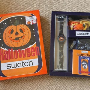Puede incluir: Un set de reloj Swatch Halloween en una caja azul oscuro, que incluye un reloj negro con cara de calabaza, patrones de estrellas y murciélagos, un accesorio de calabaza de plástico y una guía de instrucciones en el empaque naranja.