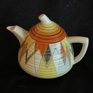 May include: A vintage ceramic teapot with a geometric design in shades of yellow, orange, brown, and white. The teapot has a lid with a ring of orange and yellow stripes.