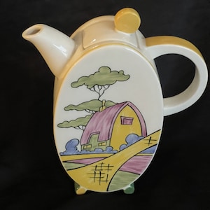 May include: A white ceramic teapot with a yellow knob on the lid. The teapot is decorated with a painted scene of a yellow house with a pink roof, green trees, and a yellow path.