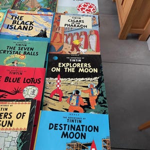 Peut inclure: Une collection de bandes dessin&eacute;es Tintin vintage, avec des couvertures color&eacute;es portant des titres tels que "L'&Eacute;toile Myst&eacute;rieuse", "On a march&eacute; sur la Lune" et "Objectif Lune". Les livres sont empil&eacute;s, pr&eacute;sentant diverses aventures du personnage embl&eacute;matique.