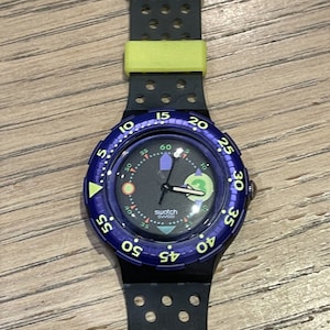 以下が含まれることがあります： 緑とオレンジの文字盤を持つ、青と黒のSwatchの腕時計。時計は、黄色のアクセントが付いた黒いゴム製のストラップをしています。