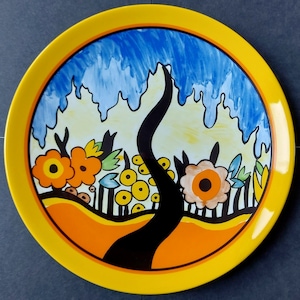 May include: A round, yellow-rimmed decorative plate with a colorful, whimsical design. The plate features a black tree, orange flowers, yellow circular shapes, and a blue sky background. The plate has a glossy finish.