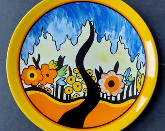 May include: A round decorative plate with a yellow rim and a vibrant, artistic design. The plate showcases a black tree, orange flowers, yellow circles, and a blue sky. The plate has a glossy finish and is likely ceramic.