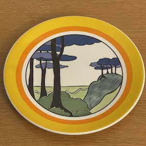 Könnte beinhalten: Ein gelber Keramikteller mit konzentrischen orangefarbenen und gelben Ringmustern. Die Mitte des Tellers zeigt eine stilisierte Landschaftsszene mit Bäumen, einem Hügel und Wasser.