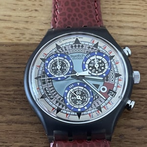 以下が含まれることがあります： パターン化された文字盤と3つのサブダイヤルを備えた、黒いSwatchクロノグラフ腕時計。赤い革バンドが付いています。文字盤には「SWATCH SWISS AG 1992」の文字があります。