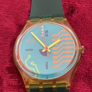 May include: A yellow and blue Swatch watch with a green band. The watch face has a circuit board design with orange and green lines. The watch has a red second hand and yellow hour and minute hands.