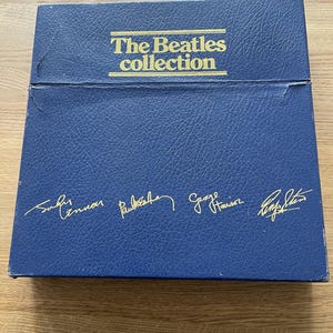 Op de afbeelding: Een blauwe boxset met de gouden tekst "The Beatles collection". De handtekeningen van John Lennon, Paul McCartney, George Harrison en Ringo Starr zijn in goud op de box gedrukt.