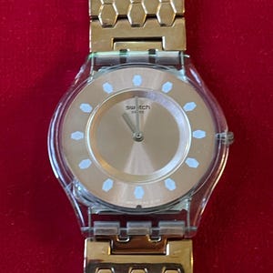 May include: A gold-toned Swatch watch with a clear plastic case and a gold dial. The watch features a honeycomb-patterned band and white hour markers. The watch is displayed against a red background.