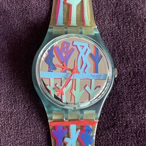 May include: A blue and green Swatch watch with a colorful abstract design on the face and band. The watch face has a silver background and the text "swatch swiss" is printed on the face.