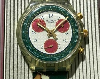 May include: A gold and white Swatch watch with a green and gold band. The watch face has a white background with black numbers and hands. The band has a repeating pattern of horses and riders. The watch face also has the words "Dressage Trot Jump" and "Swatch Riding Team" printed on it.