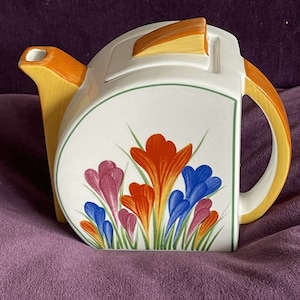 Könnte beinhalten: Eine weiße Teekanne aus Keramik mit einem gelben und orangefarbenen Henkel und Deckel. Die Teekanne zeigt ein Blumenmuster aus blauen, orangefarbenen, rosa und gelben Krokusblüten.