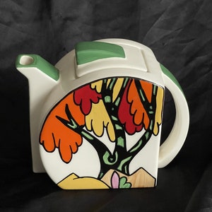 May include: White ceramic teapot with a stylized tree design in shades of orange, yellow, green, and black. The teapot has a green lid and a handle.
