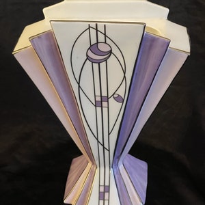 Könnte beinhalten: Eine weiße Keramikvase mit einem geometrischen Design in Violett- und Goldtönen. Die Vase hat eine breite, ausgestellte Öffnung und einen schmalen Boden.