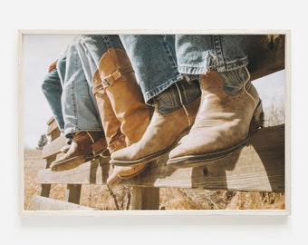May include: Three pairs of brown cowboy boots with worn leather and stitching details. The boots are worn by people sitting on a wooden fence.
