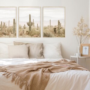May include: Three framed prints of a desert landscape with cacti and mountains in muted tones of brown and green. The prints are hanging on a white wall above a bed with white linens and a brown throw blanket.