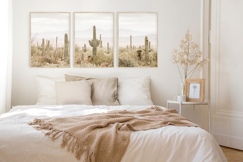 May include: Three framed prints of a desert landscape with cacti and mountains in muted tones of brown and green. The prints are hanging on a white wall above a bed with white linens and a brown throw blanket.