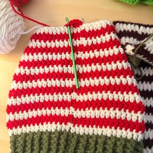 May include: A partially completed crochet hat with red and white stripes, a green brim, and a green crochet hook. Red and white yarn balls are in the background. The hat is handmade.