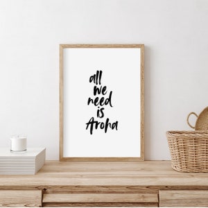 Può includere: Una stampa incorniciata con sfondo bianco e testo nero che recita "all we need is aroha". La stampa è appoggiata su un tavolo di legno con una candela e un cesto di vimini.