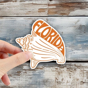 May include: A sticker featuring a seashell design with the word "FLORIDA" in orange. The sticker is held by a hand against a weathered, blue-toned wooden background. The Planner Family logo is in the bottom right corner.