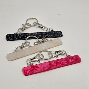 Puede incluir: Tres llaveros con colgantes rectangulares. Uno es negro con motas blancas, otro es blanquecino y el tercero es rosa vibrante. Cada llavero tiene un cierre plateado y un llavero.