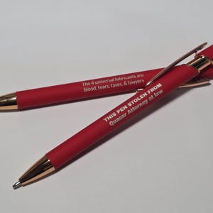 May include: Two red ballpoint pens with gold-colored accents and stylus tips. One pen features the text "The 4 universal lubricants are blood, tears, taxes, & lawyers" and the other reads "THIS PEN STOLEN FROM Quasar Attorney at law" in white.