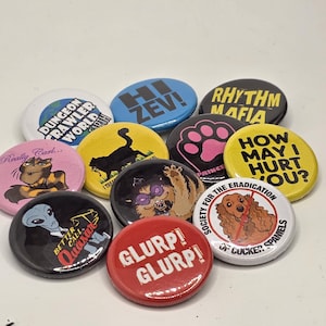 May include: A selection of circular badges with diverse designs and text. The badges display phrases such as "How May I Hurt You?" and "Better Call Quasar." Other designs include a cat silhouette, a paw print, and a dog illustration.