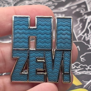 May include: A rectangular pin with the word "HI ZEV!" in a bold, stylized font. The letters are filled with a wavy blue pattern and outlined in silver. The exclamation point is also silver.