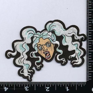 May include: An embroidered patch featuring a character with pale blue, wavy pigtails and a tan face with freckles, wide eyes, and an open mouth showing fangs. The patch has a black border and is positioned next to a ruler showing 10 cm in width.