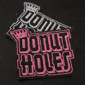May include: Two embroidered patches with the words "DONUT HOLES" in black block letters with a pink outline. Each patch has a crown above the text. One patch is pink and black, and the other is white and black.