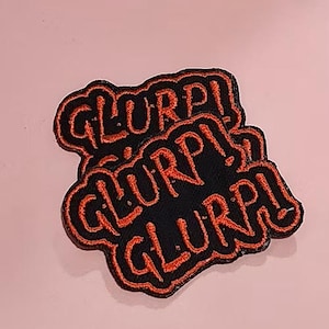 May include: Two black embroidered patches with the word "GLURP!" in orange stitching. The patches are stacked on top of each other against a light pink background. The patches have an orange border.