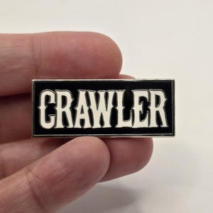 May include: A rectangular enamel pin with the word "CRAWLER" in bold white letters on a black background. The pin has a silver border and is held in a person's hand.