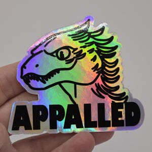 May include: A holographic sticker featuring a stylized dragon head with the word "APPALLED" in bold black letters. The sticker has a rainbow iridescent finish, with black outlines and details.