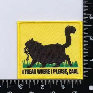 May include: Embroidered patch with a yellow background and a black cat silhouette carrying a snake. The patch reads "I TREAD WHERE I PLEASE, CARL" in white text. The cat is standing on green grass.