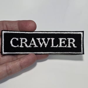 May include: Black rectangular patch with the word "CRAWLER" embroidered in white, outlined with a white border. The patch is likely for clothing or accessories.