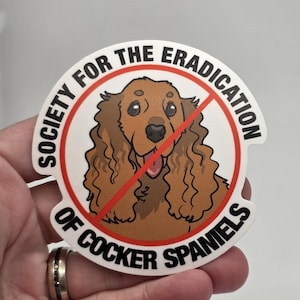May include: A round sticker with the text "SOCIETY FOR THE ERADICATION OF COCKER SPANIELS" in black, surrounding a cartoon Cocker Spaniel image with a red slash through it. The dog is brown with a pink tongue.
