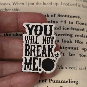 Puede incluir: Pin de esmalte en blanco y negro con el texto "YOU WILL NOT BREAK ME!" y un gráfico de bomba. El pin es rectangular con fondo blanco y letras negras. El pin está en la mano de una persona.