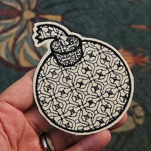 May include: A circular patch with a black and white geometric pattern. The patch has a cream-colored background and a black border. A smaller, detailed design is at the top. The patch is held in a hand.