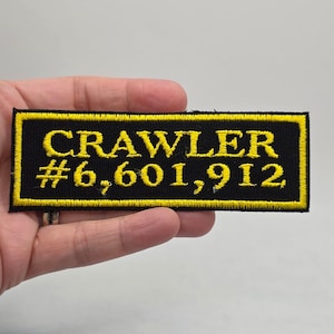 May include: Black rectangular patch with yellow embroidered text that reads "CRAWLER #6,601,912". The patch has a yellow border and is held in a hand against a neutral background. The patch is likely for a jacket or bag.