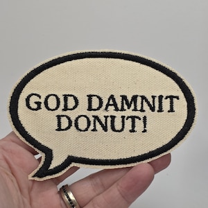 Könnte beinhalten: Ein beigefarbener Aufnäher in Form einer Sprechblase mit dem Text "GOD DAMNIT DONUT!" in schwarzen Buchstaben. Der Aufnäher hat einen schwarzen Rand und wird von einer Hand gehalten.