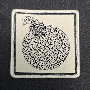 May include: A square embroidered patch featuring a cream-colored background with a black border. The center displays a circular graphic of a bomb with a lit fuse, filled with a repeating geometric lattice pattern in black thread embroidery.