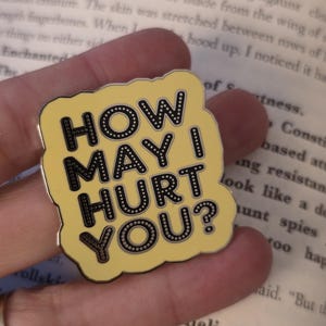May include: A yellow enamel pin with the text "HOW MAY I HURT YOU?" in black, outlined in white dots. The pin is held against a backdrop of a book's pages.