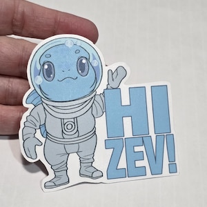 May include: A sticker featuring a cartoon character in a spacesuit with a bubble helmet. The character is light blue with large eyes and a smiling mouth. The text "HI ZEV!" is in blue block letters.