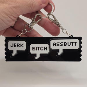 Könnte beinhalten: Schwarzer Schlüsselanhänger mit drei weißen, verpixelten Sprechblasen. Die Sprechblasen enthalten die Wörter "JERK", "BITCH" und "ASSBUTT". Der Schlüsselanhänger hat silberfarbene Verschlüsse und einen Schlüsselring.