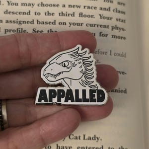 May include: A silver-colored enamel pin featuring a stylized dragon head with the word "APPALLED" in bold, black letters. The pin is set against the backdrop of an open book.