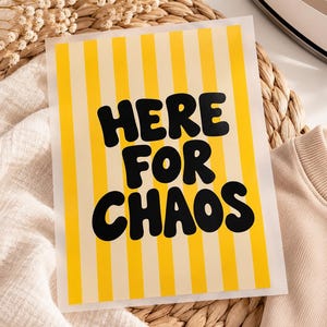 Könnte beinhalten: Ein rechteckiger Bügeltransferbogen mit vertikalen gelben und cremefarbenen Streifen und dem fetten schwarzen Text HERE FOR CHAOS, der in einer abgerundeten, blasenartigen Schriftart in der Mitte des Designs zum Aufbügeln gedruckt ist.