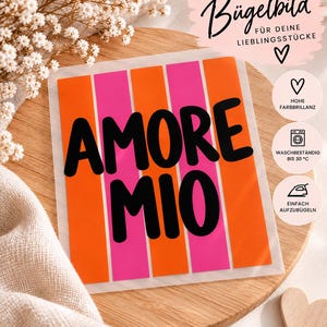 Könnte beinhalten: Ein quadratisches Bügelbild mit dem Text AMORE MIO in fetten schwarzen Buchstaben auf einem Hintergrund aus abwechselnden vertikalen orangefarbenen und pinken Streifen. Das Design enthält kleine runde Symbole mit Texten zu hoher Farbbrillanz, Waschbeständigkeit bis 30 Grad Celsius und einfacher Anwendung.