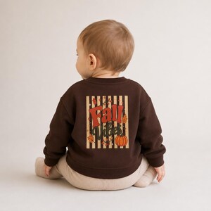 Könnte beinhalten: Ein dunkelbraunes Langarm-Sweatshirt mit einem Grafikdruck auf der Rückseite, der vertikale beige Streifen, orangefarbene und rote Herbstblätter, einen Kürbis und den Schriftzug Fall Vibes in einer Retro-Schriftart zeigt. Gerippter Rundhalsausschnitt.