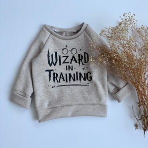 Può includere: Una maglietta a maniche lunghe in maglia a nido d'ape beige con una stampa grafica nera che dice "Wizard in Training" con un paio di occhiali e una bacchetta.