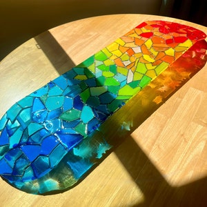 Rainbow stained glass skateboard deck artwork on table in sunlight casting colorful shadow.