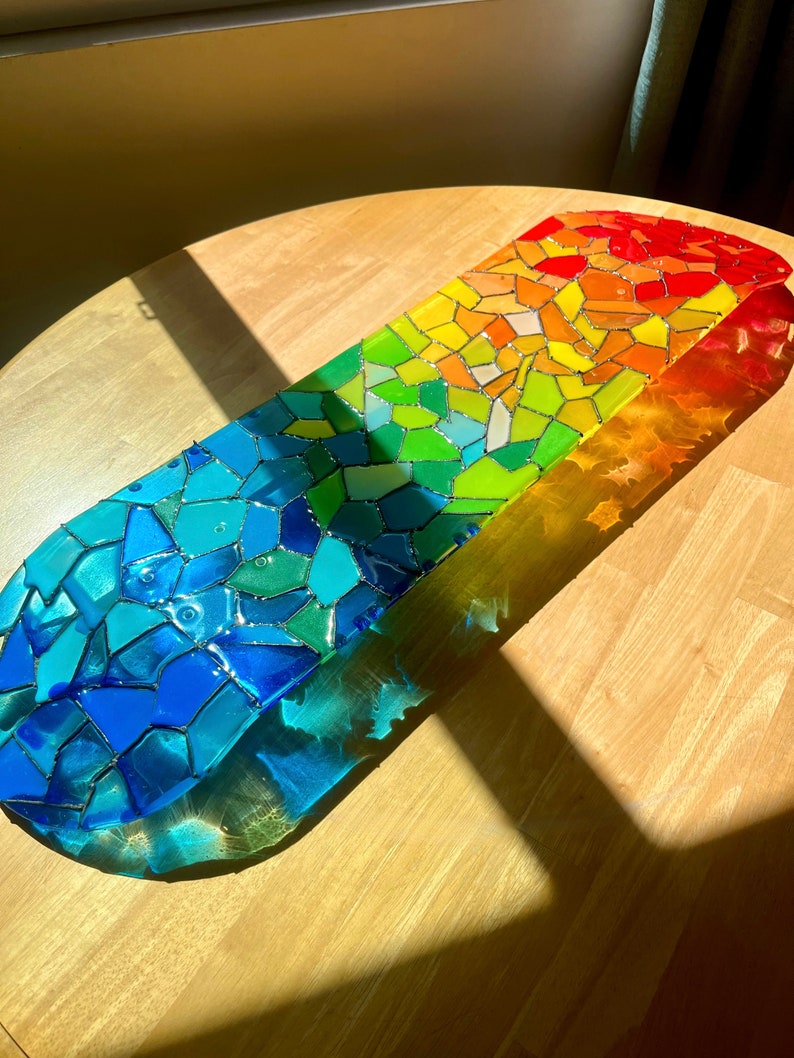 Rainbow stained glass skateboard deck artwork on table in sunlight casting colorful shadow.